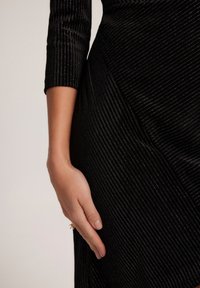 Czarna sukienka z prążkowanego materiału o teksturowanej powierzchni, z rękawami o długości trzy czwarte i asymetrycznym krojem, z ręką opartą na boku.