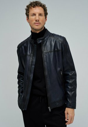 Hombre con cabello rizado que lleva una chaqueta de cuero negra sobre un suéter de cuello alto negro, posando frente a un fondo liso de color gris claro.