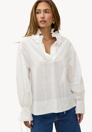 Femme portant un chemisier blanc à manches longues avec un col à volants et un jean bleu, regardant sur le côté, avec des boucles d'oreilles en or et des bagues visibles.