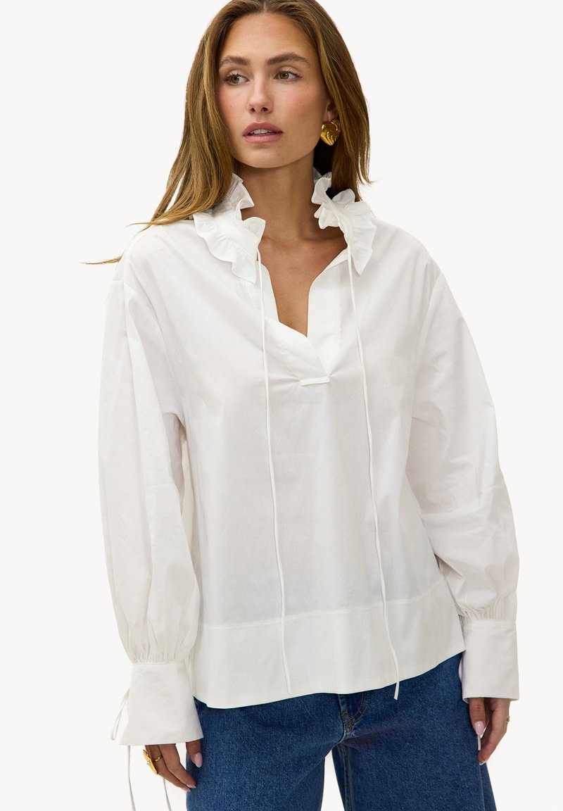 Femme portant un chemisier blanc à manches longues avec un col à volants et un jean bleu, regardant sur le côté, avec des boucles d'oreilles en or et des bagues visibles.