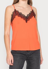 Oranje hemdje met dunne bandjes, voorzien van een zwarte kanten V-hals en een gladde textuur. Gecombineerd met donkere spijkerbroek.