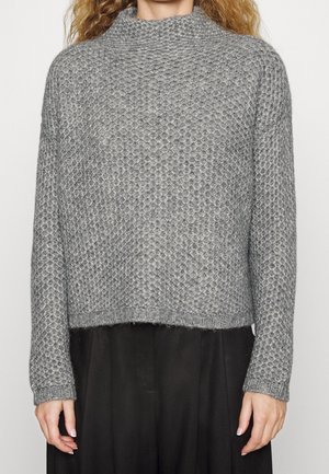 Šedý hrubý pletený rolák s texturovaným vzorem nošený přes černé kalhoty, zobrazující trup a paže osoby s kudrnatými vlasy.