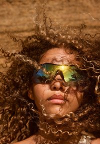 Occhiali da sole sportivi con una montatura scura e lenti riflettenti che mostrano una sfumatura di verde e oro. Capelli ricci e crespi parzialmente coprono il volto.