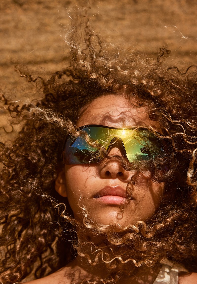 Occhiali da sole sportivi con una montatura scura e lenti riflettenti che mostrano una sfumatura di verde e oro. Capelli ricci e crespi parzialmente coprono il volto.