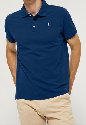 Mann trägt ein dunkelblaues Poloshirt mit kleinem weißen Golfer-Logo und beiger Hose, steht mit den Händen hinter dem Rücken.