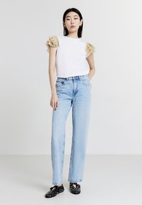 Maglietta bianca a maniche corte con dettagli beige arricciati sulle spalle, abbinata a jeans a gamba larga azzurri e scarpe nere con fantasia.