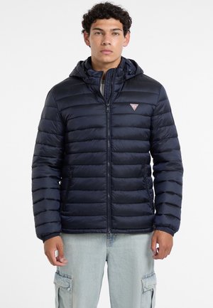 Joven con chaqueta acolchada azul marino con capucha y pantalones vaqueros cargo azul claro, de pie frente a un fondo blanco liso.