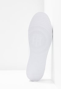 Suela de zapatilla blanca con patrones de agarre texturizados, puntos circulares y detalle de logo en relieve; el material suave se transita hacia el borde perimetral.