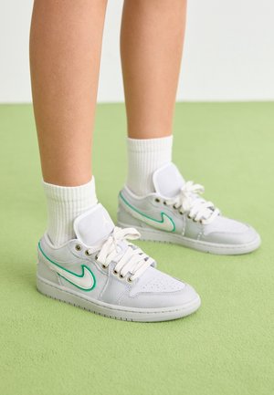 Ben iført hvide sneakers med turkis Nike-swoosh og hvide ribstrikkede sokker, stående på lys grøn gulv.