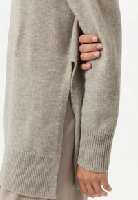 Ljusgrå stickad tröja med ribbade muddar och sidoflikar, med en mjuk textur och avslappnad passform. Hand synlig på sidan.