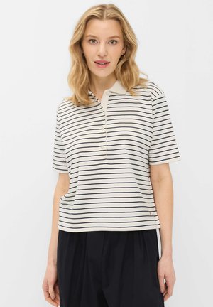 Lyshåret kvinde iført hvid kortærmet polo med tynde sorte horisontale striber og sorte plisserede bukser mod hvid baggrund.