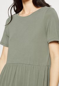 Olivově zelené šaty s kulatým výstřihem a krátkými rukávy. Mají přiléhavé tělo a rozšířenou, nařasenou sukni pro uvolněný střih.