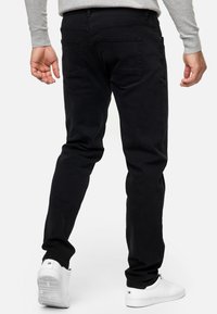 Man die een zwarte spijkerbroek, witte sneakers en een lichtgrijs shirt met lange mouwen draagt, gezien van achteren tegen een witte achtergrond.