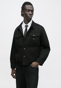 Schwarze Jeansjacke mit Kragen, Fronttaschen und silbernen Beschlägen. Kombiniert mit einem weiß gestreiften Hemd und einer Krawatte. Strukturierter Stoff.