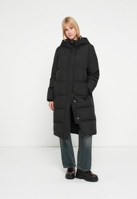 Must kapuutsiga pufferjakk, millel on nööbitav eesmine osa, tikkimismustriga ja pikad varrukad, kombineeritud lahtiste teksapükste ja massiivsete saabastega.