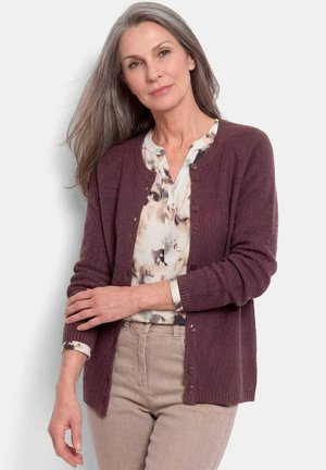 Femme mûre aux longs cheveux gris porte un cardigan bordeaux sur une blouse à motifs et un pantalon beige, debout avec un bras croisé.