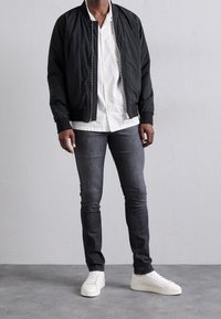 Svart bomberjacka, vit skjorta med knappar, grå slim-fit jeans och vita sneakers. Jackan har dragkedjestängning och ribbade ärmslut.