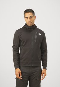 Uomo che indossa una felpa nera con zip e cappuccio, con una tasca sul petto che presenta il logo The North Face, in piedi contro uno sfondo neutro.