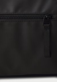 Sac en tissu noir avec une surface texturée, une fermeture éclair proéminente avec un tirette noir, et une sangle de transport tissée pour plus de fonctionnalité.