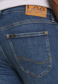 Vaqueros de mezclilla en azul oscuro con una etiqueta de cuero que muestra "Lee". Presenta costuras amarillas y diseño en el bolsillo trasero. Tejido texturizado.