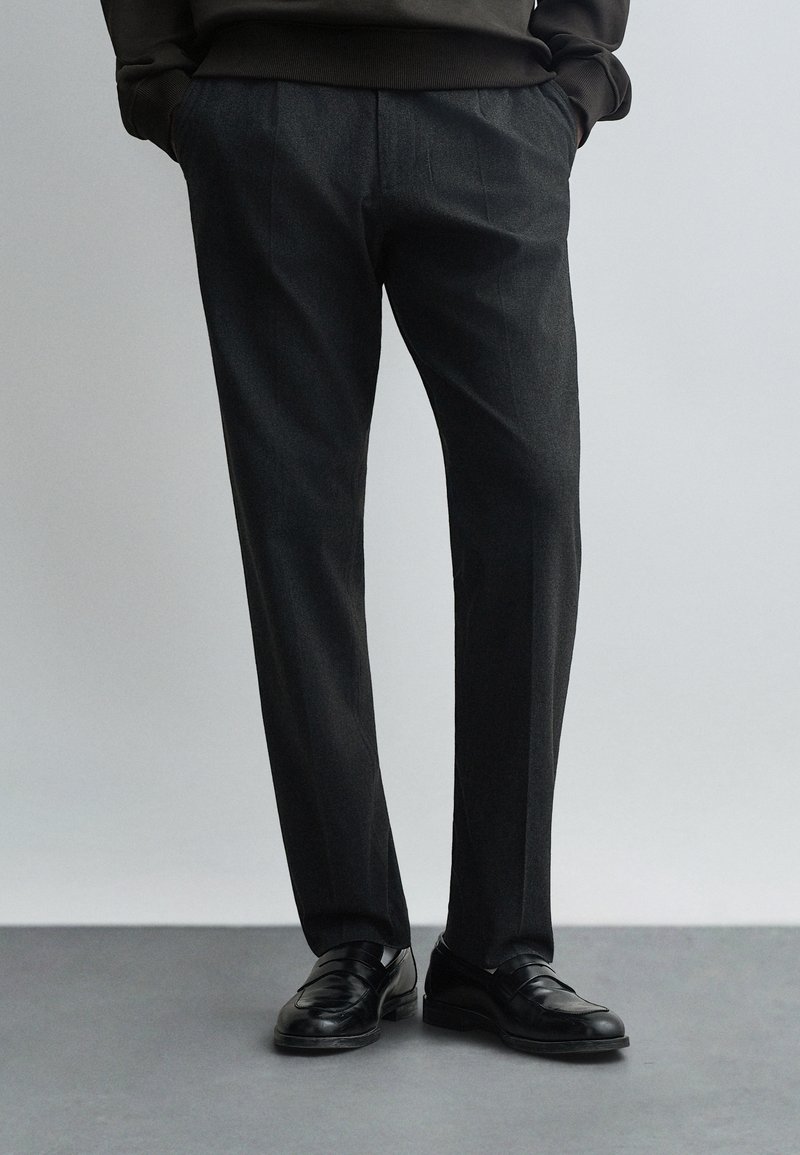 Homme portant un pantalon gris foncé taille slim, mains dans les poches, et des mocassins en cuir noir, debout sur un sol gris.