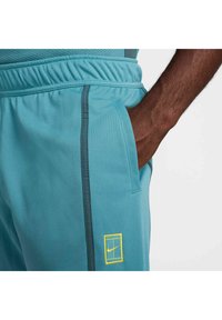 Turkise Nike treningbukser med elastisk midjebånd, sidelommer og grå detaljer. Har en gul logo på nederste del av benet.