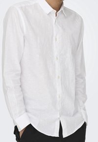Camisa blanca de lino con botones y mangas largas, con un cuello clásico y una tela ligeramente texturizada. Los botones son pequeños y de color claro.