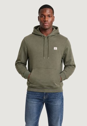 Mand iført olivengrøn hættetrøje med forlommer og lille logo på brystet, kombineret med blå jeans, står mod en ensfarvet baggrund.