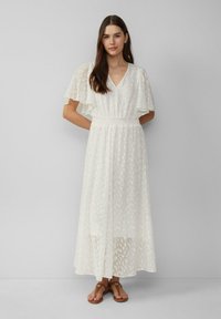 Donna con lunghi capelli castani, vestita con un maxi abito bianco a fantasia con maniche a sbuffo e sandali marroni, in piedi davanti a uno sfondo grigio semplice.
