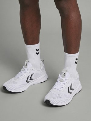 Nogi w białych butach sportowych z czarnymi logo w kształcie zygzaka oraz pasujące białe skarpetki za kostkę z czarnymi zygzakami na szarym tle.