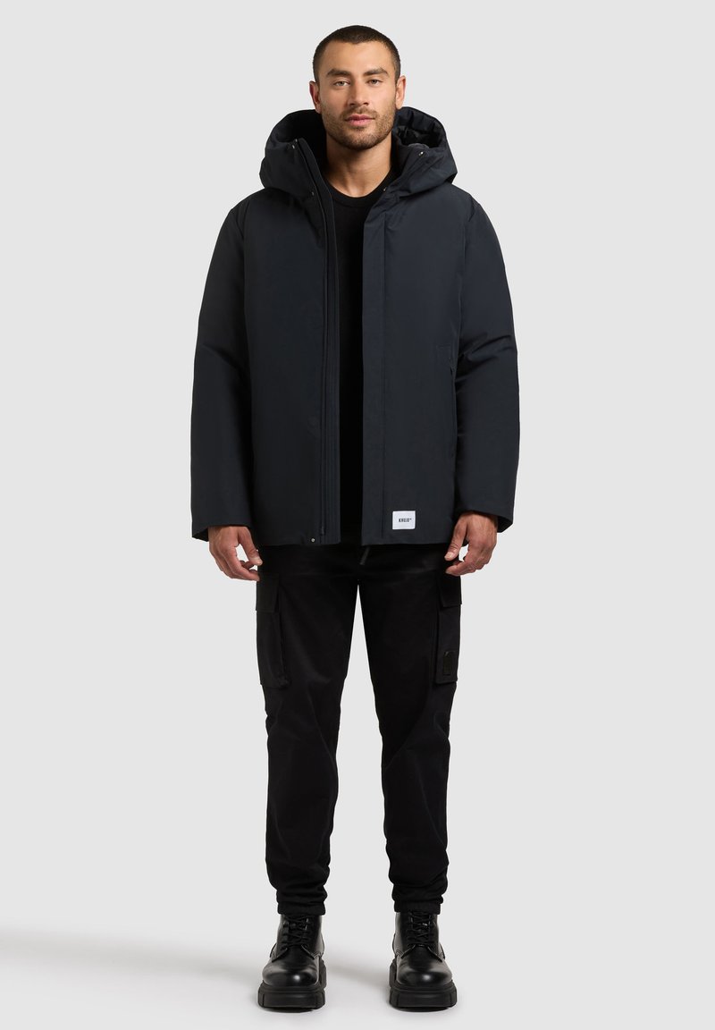 Black insulated jacket with a hood, zip front, and side pockets, worn with black cargo pants and black lace-up boots.