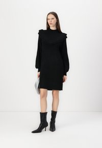 Juoda megzta suknelė su plačia raukšlėta apykakle, ilgomis pūstomis rankovėmis ir tiesia siluetu; derinama su juodais aulinukais ir sidabrine rankine.