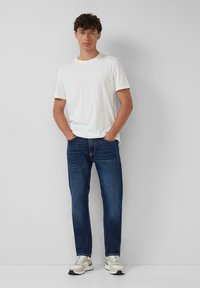 Witte t-shirt met korte mouwen, gestoken in donkerblauwe denim jeans, gecombineerd met lichtgekleurde sneakers. Eenvoudige achtergrond.