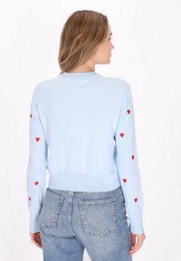 Ljusblå cardigan med röda hjärtan på ärmarna, ribbade muddar och fåll, mjukt stickat material, figursydd design. Avslappnad stil.
