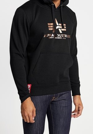 Mężczyzna w czarnej bluzie z kapturem Alpha Industries z złotym logo i ciemnoniebieskich dżinsach na białym tle.