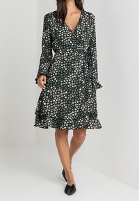 Grünes und cremefarbenes Wickelkleid mit Sternmuster, langen Ärmeln und rüschenbesäumtem Saum. Hergestellt aus leichtem Stoff, ausgestattet mit einem Bindegürtel.