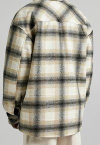 Chaqueta de franela a cuadros en tonos beige, negro y blanco, vista desde atrás, con mangas largas y puños abotonados.