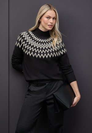 Maglione nero a maglia con motivo zigzag bianco sul giromanica, colletto alto, abbinato a pantaloni neri strutturati e una borsa a tracolla nera.