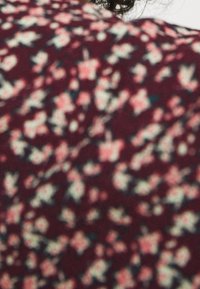 Cheveux bouclés et foncés partiellement visibles au-dessus d'un tissu bordeaux à motif floral blanc et rose.