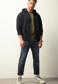 Felpa con zip di colore blu navy con tasca frontale, t-shirt verde oliva sotto, jeans blu scuro e sneaker bianche con dettagli verdi.