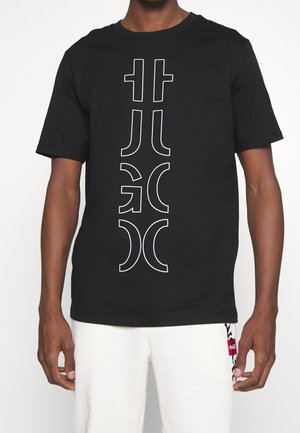 Man wearing a black T-shirt with a vertical white abstract design and white pants with a small red and black patterned tag.