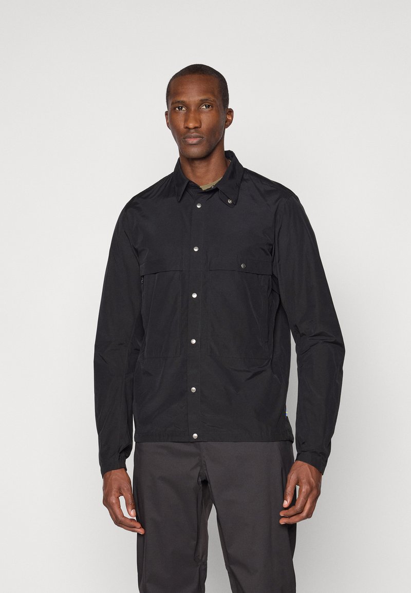 Black lightweight jacket featuring snap-button closure, two front pockets, and a pointed collar. Smooth texture with a minimalistic design.