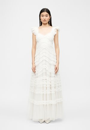 Nő fehér, földig érő, ujjatlan ruhában, fodros rétegekkel és tüll anyaggal, világos, egyszínű háttér előtt állva.