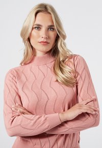 Różowy sweter wykonany na drutach z wysokim kołnierzem, z falistym wzorem tekstury i ściągaczami na rękawach. Miękka tkanina z subtelnymi detalami.