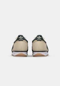 Adidas sneakers met een beige suede bovenkant, zwarte mesh zijkanten, groene hielaccenten, witte tussenzool en een gumrubberen buitenzool.