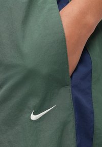 Αντιανεμικό Nike σε σκούρο πράσινο, με λογότυπο σε ανοιχτό γκρι. Το υλικό φαίνετ αι ελαφρύ και με υφή, με αντίθετη σκούρα μπλε επένδυση στο εσωτερικό.