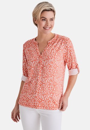 Frau mit kurzen blonden Haaren, die ein korallenrotes und weiß gemustertes langärmliges Hemd und eine weiße Hose trägt, lächelt und schaut nach rechts.