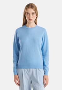 Junge Frau mit hellbraunem Haar trägt einen hellblauen Rundhalspullover und hellblaue gestreifte Hosen und steht vor einem weißen Hintergrund.