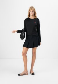 Vrouw die staat en een zwart langemouwen top met kleine rode details draagt, een zwarte geplooide rok, beige en zwarte ballerina's, terwijl ze een zwarte handtas vasthoudt.