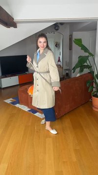Femme en trench-coat beige attachant la ceinture dans un salon moderne avec un sol en bois, un canapé orange, une grande plante et un meuble TV.